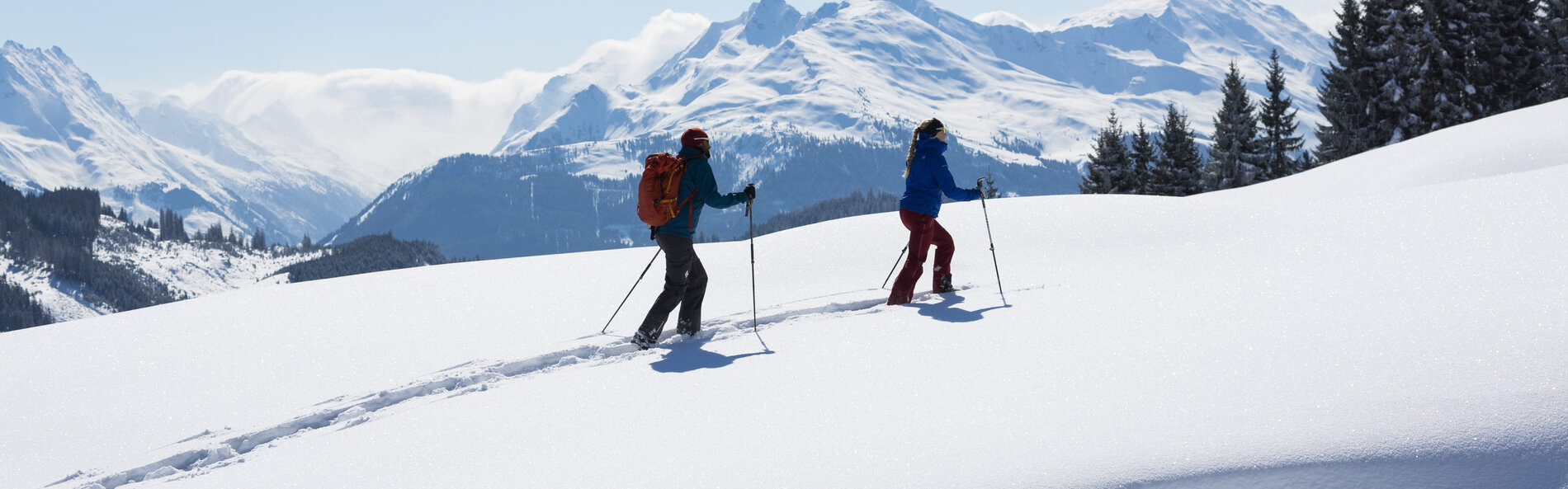 Eine Frau und ein Mann gehen hintereinander mit den Schneeschuhen einen verschneiten flachen Hang bergauf. Im Hintergrund sieht man tief verschneite Berge. Der Himmel ist blau und die Sonne scheint. 