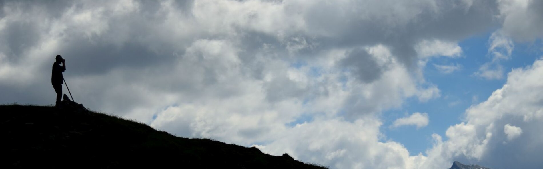 Ein Bauer steht auf eine, Berg mit einem Stock. Ma sieht nur die Silhouette von Berg und Bauer. Im Hintergrund ziehen weiße Wolken vorbei.