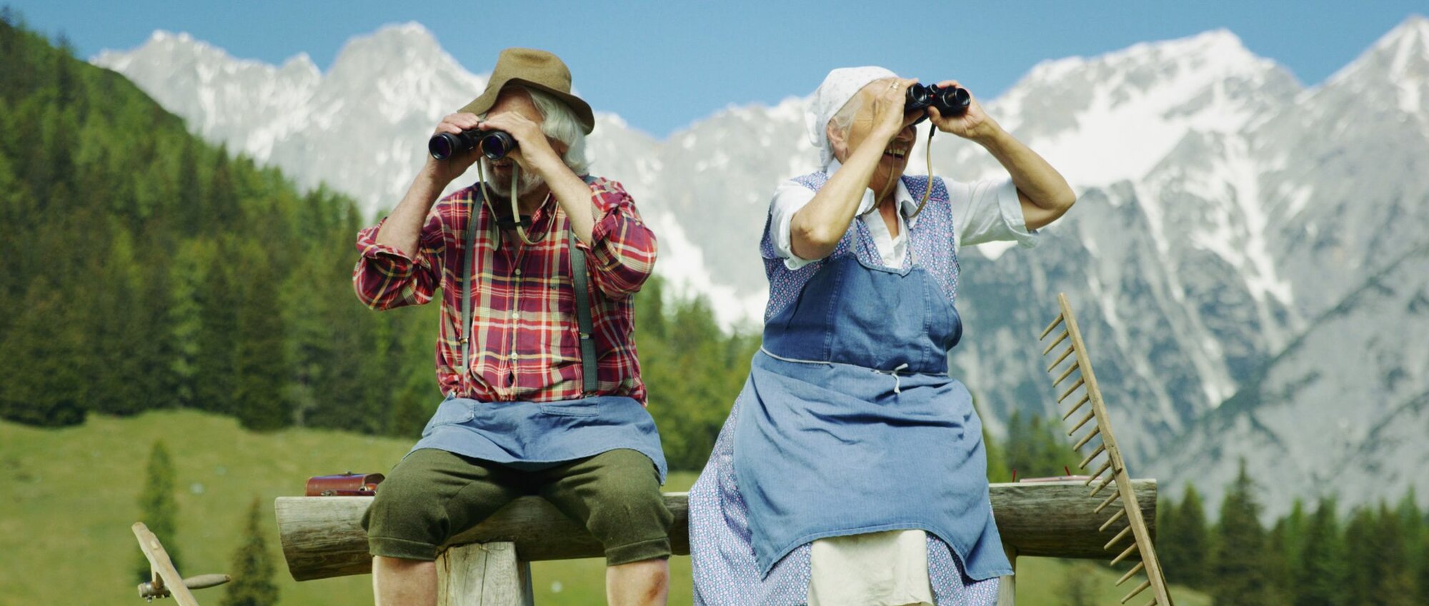 Eine ältere Bäuerin und Bauer sitzen mit Fernglässer in der Hand auf einer Bank. Sie schauen in die Ferne. Im Hintergrund Berge und Wald.