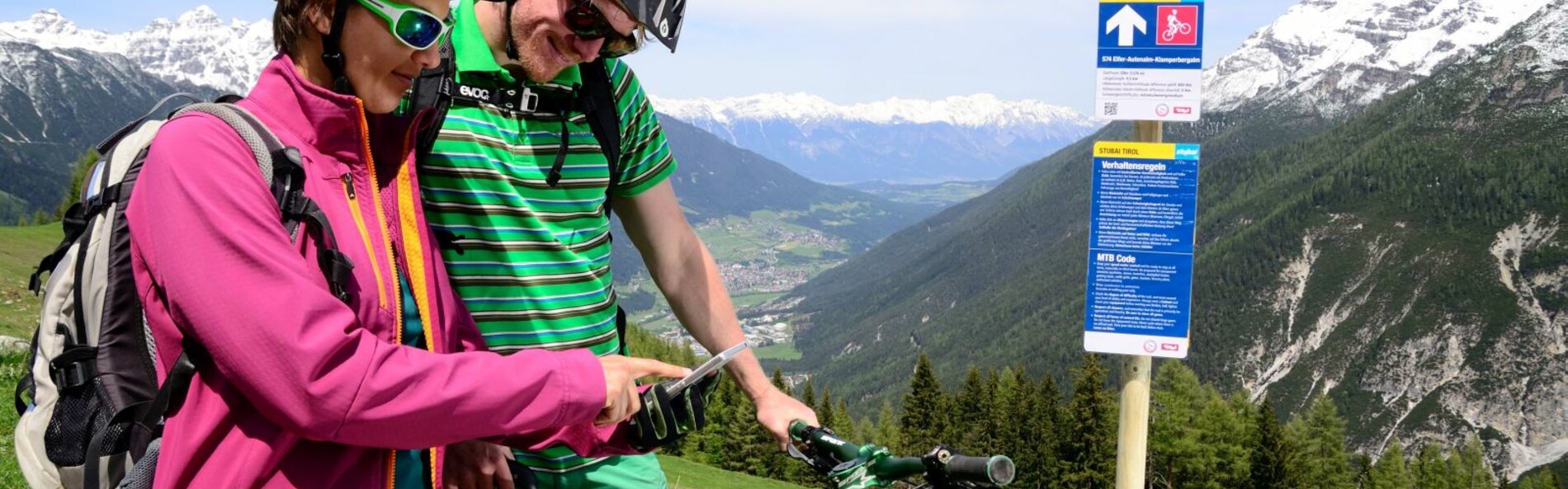 Zwei Mountainbiker versuchen sich zu orientieren und betrachten ihr Smartphone. Dabneben steht ein Mountainbikeroutenschild, ein blaues Schild mit den Verhaltenregeln und einem Richtungspfeil.