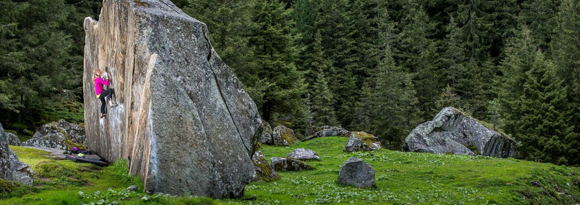 In einer Almwiese steht ein großer Felsblock, an dem eine junge Frau klettert. Im Hintergrund Fischtenwald.