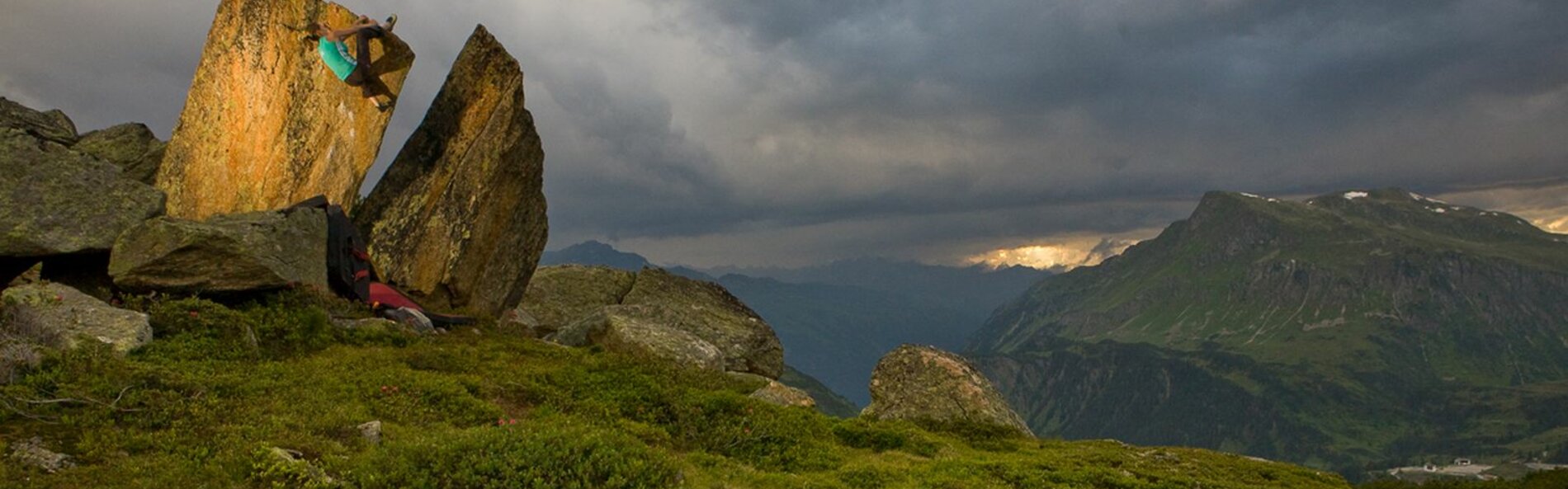 Felsblock steht in einer Wiese. Eine Frau klettert daran hinauf. Im Hintergrund ein Tal und Berge.