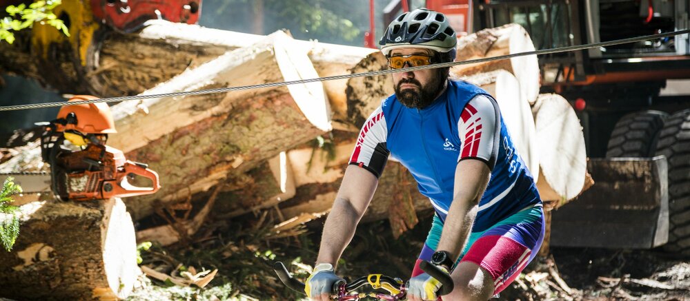 Mountainbikefahrer fährt mit erschrockenen Gesicht auf ein Stahlseil zu, welches auf Kopfhöhe gespannt ist. Im Hintergrund sind forstarbeiten am Gange