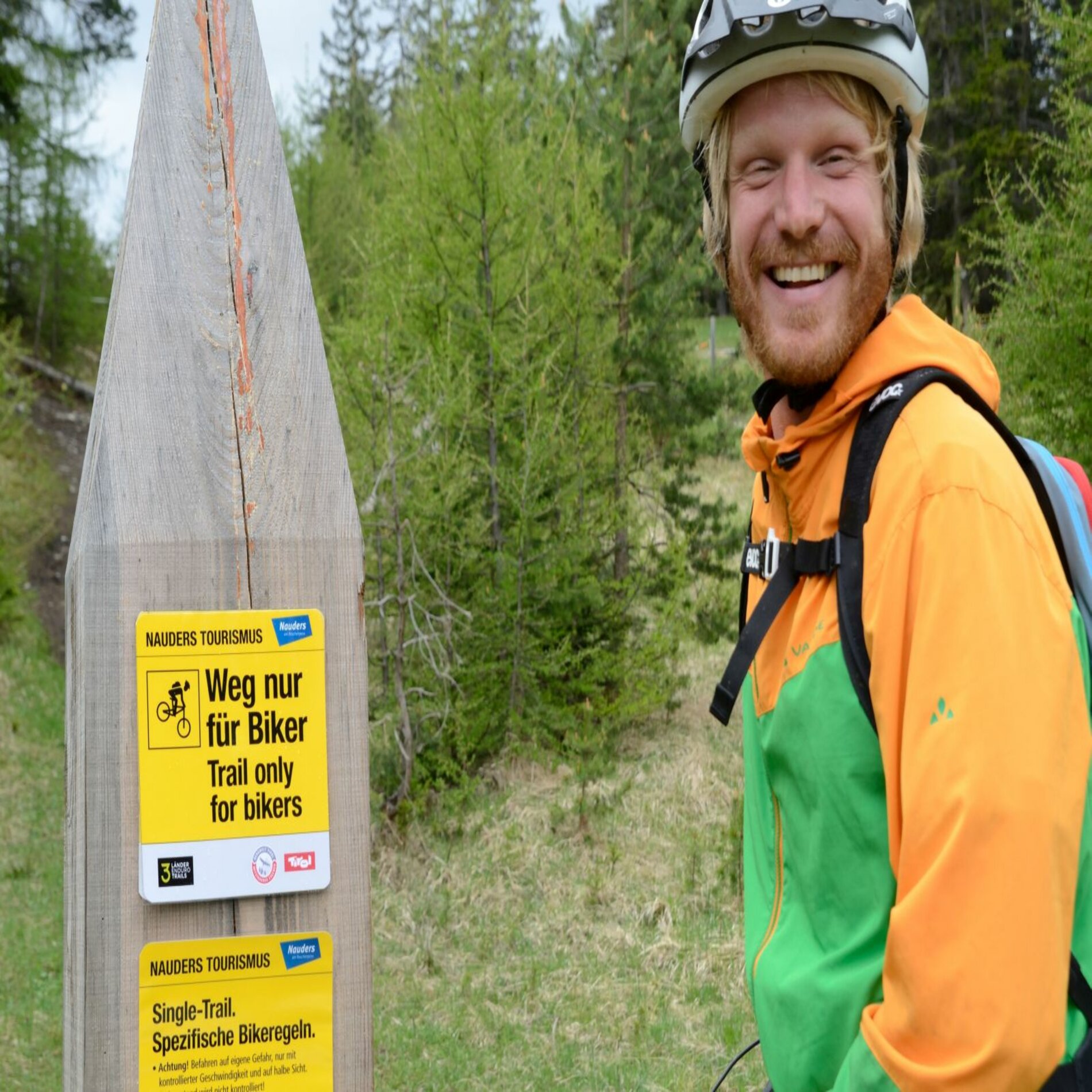 Ein Mountainbiker steht neben einem Singletrailschild und lächelt.