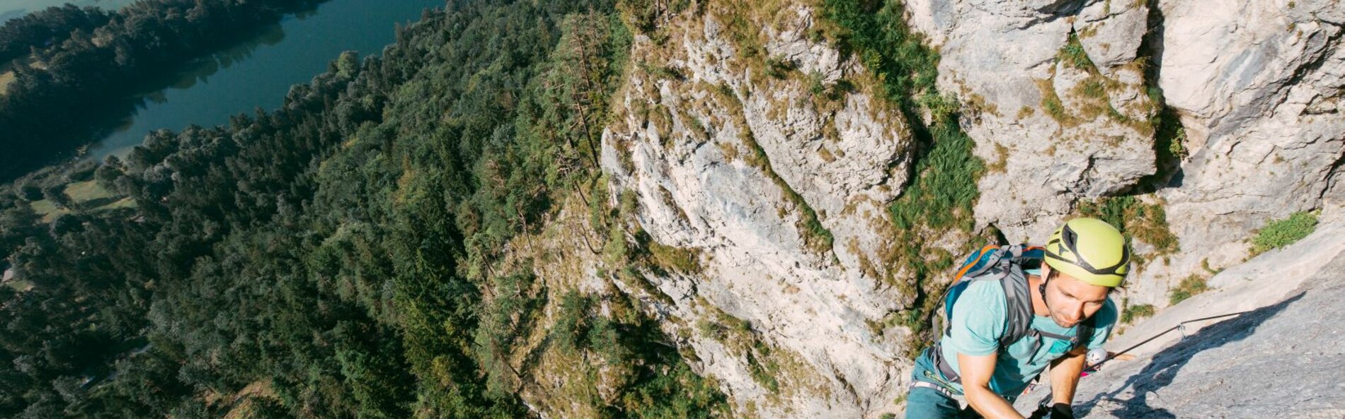 Ein juner Mann klettert entlang eines Drahtseils. Im Hintergrund sieht man die Felswand und im Tal Wald mit einem See.