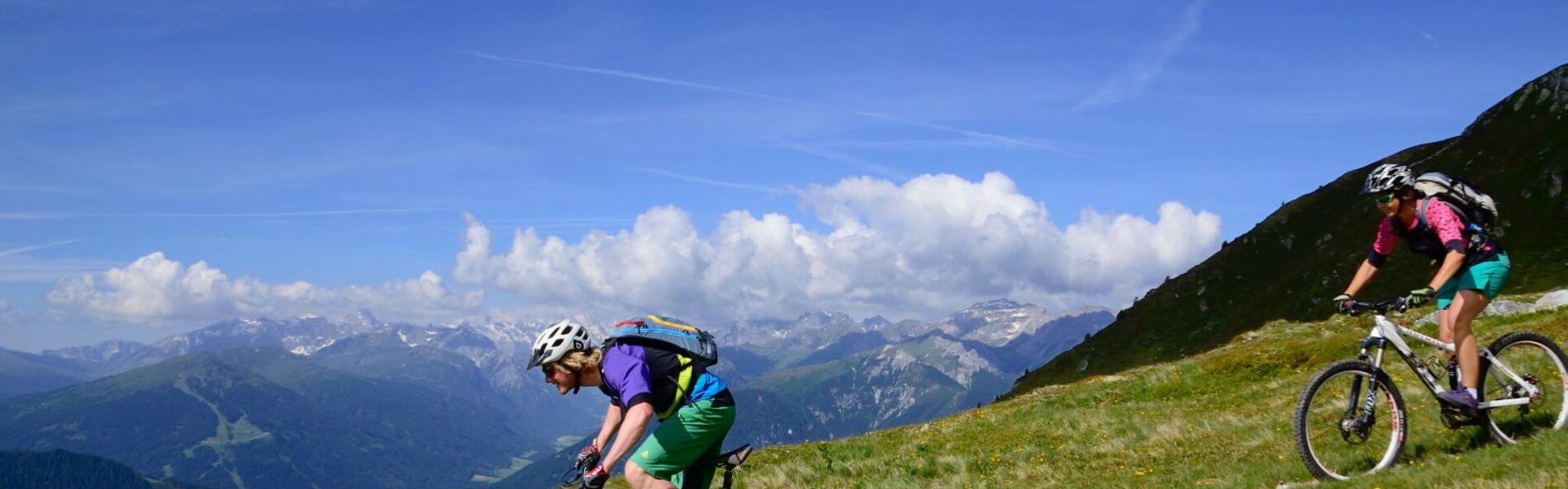 Zwei Mountainbiker auf einem Singletrail. Sie befahren den Trail abwärts, im Hintergrund blauer Himmel mit einigen Wolken am Horizont.