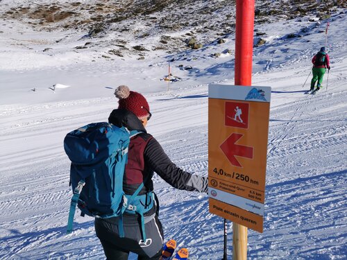 Skitourengeherin quert die Piste mit großem Abstand zum Vordermann, im Vordergrund ein Richtungsschild des Pistentouren Leitsystems mit Zusatzschild: Piste einzeln queren