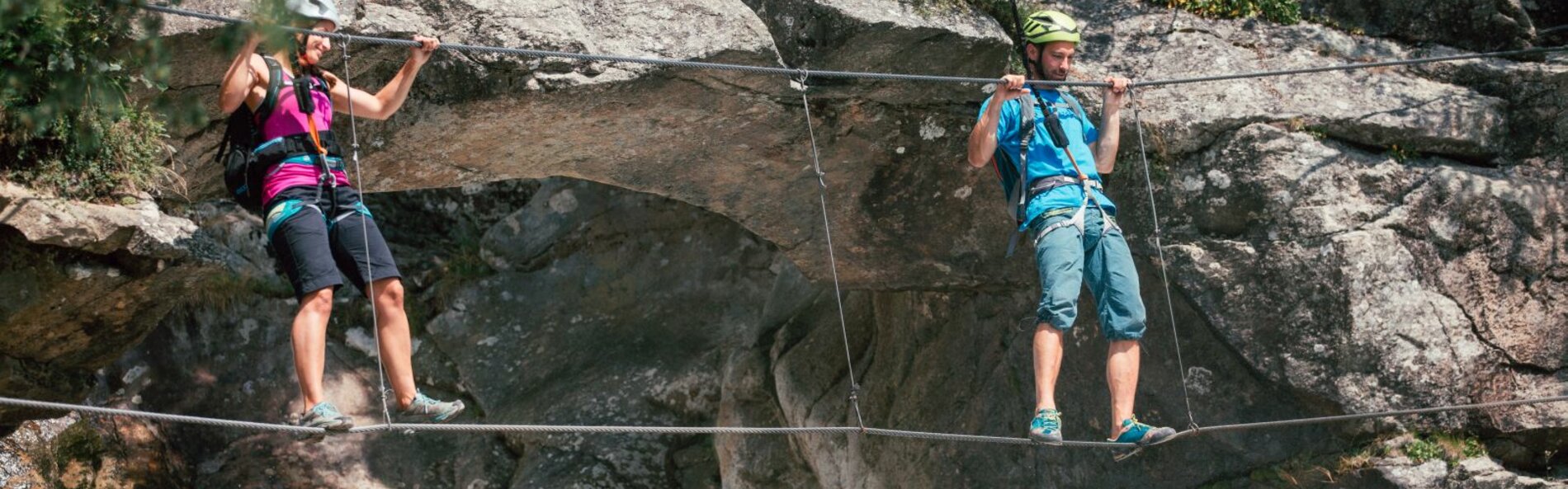 Two climbers are crossing a steel rope bridge. A rock face can be seen in the background.