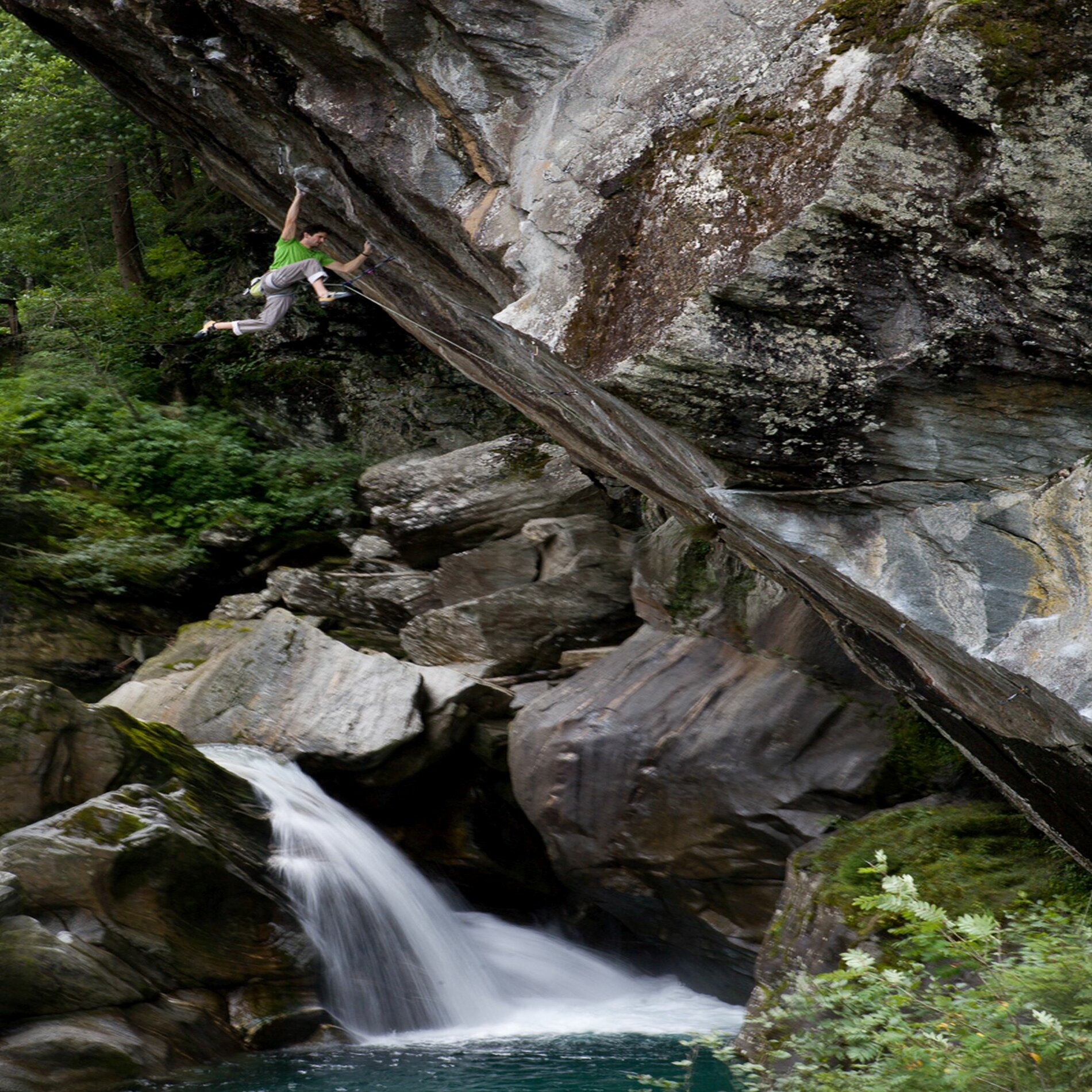 Ein Mann klettert eine steile schräge Wand hinauf. Im Hintergrund Wald und ein Bach der durch Felsblöcke fließt. 