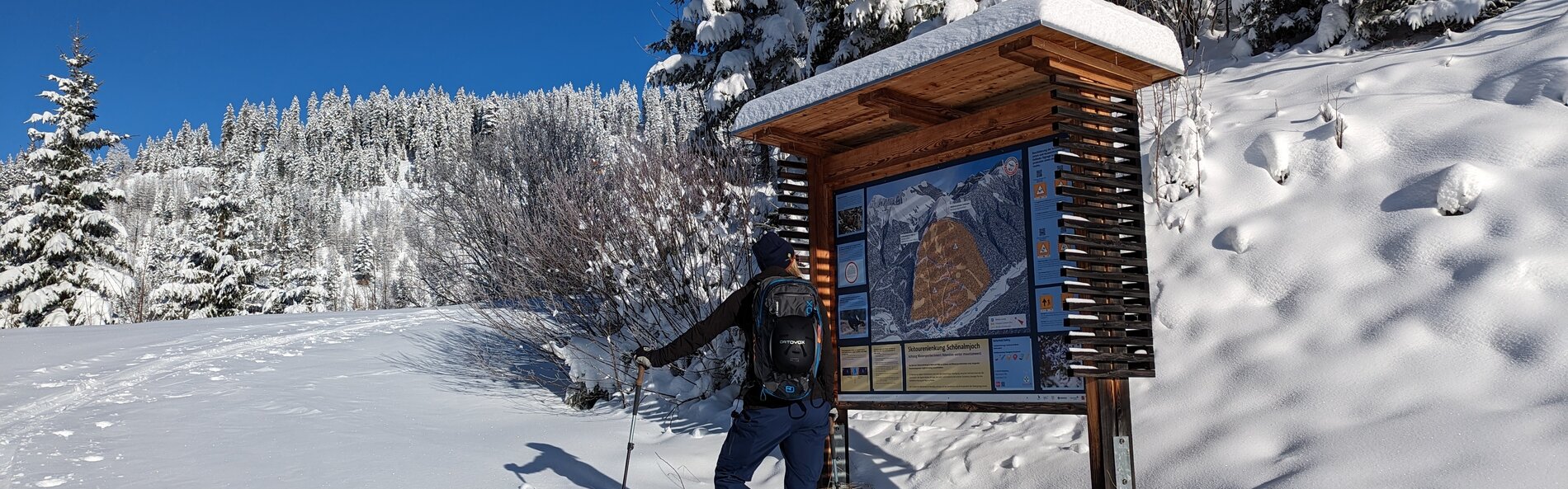 Tief verschneite Landschaft mit eingeschneiten Bäumen, im Vordergrund betrachtet eine Skitourengeherin die Skitouren Infotafel.