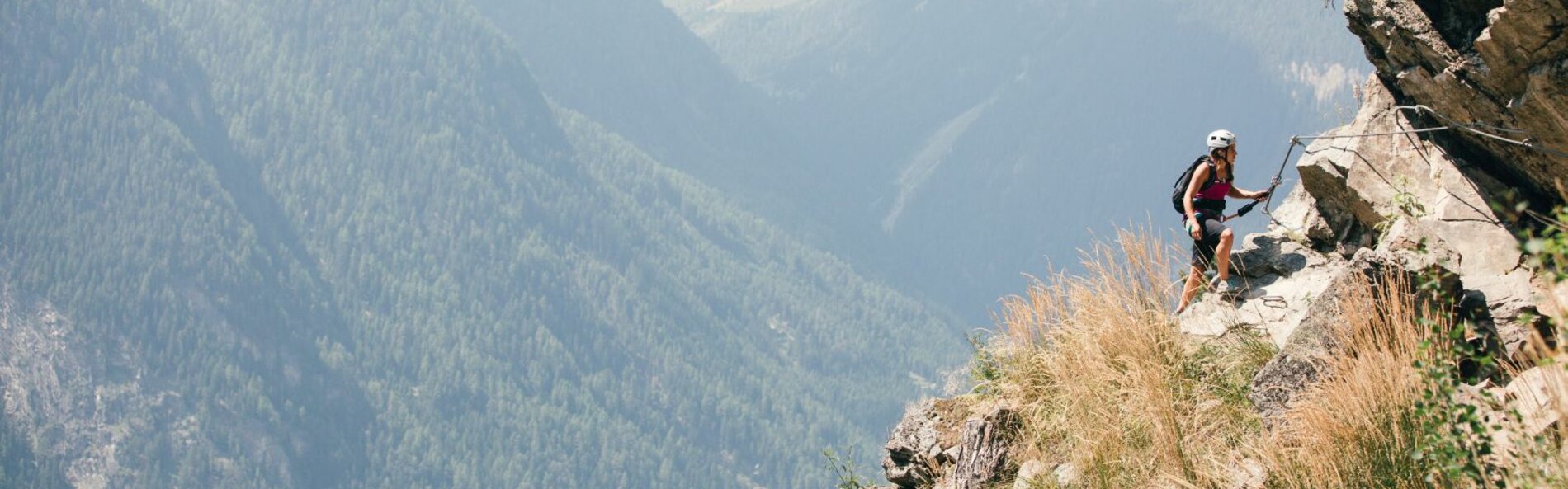 Eine Frau kletten in Klettersteigausrüstung entlang eines Drahtseil eine bewachsene Felswand entlang. Im Hintergrund sind bewaldete Berghänge und Täler.