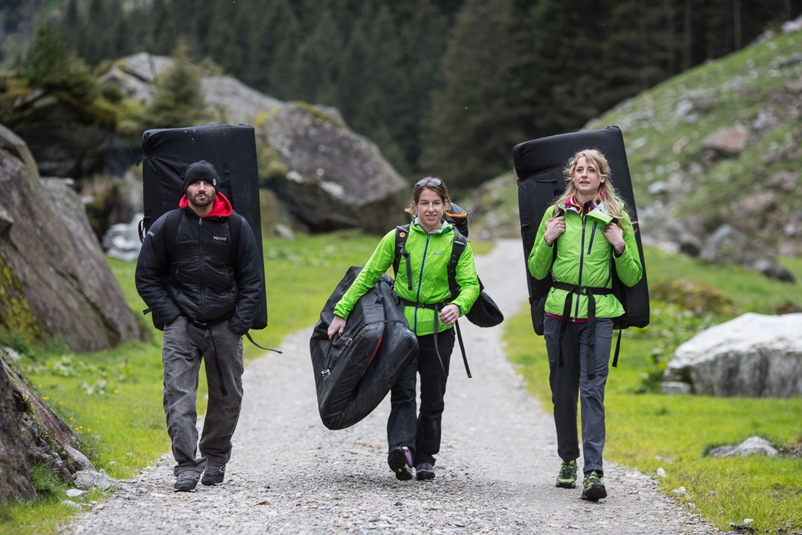 Drei Personen mit dicken Jacken und Bouldermatten gehen auf einer Forststraße.