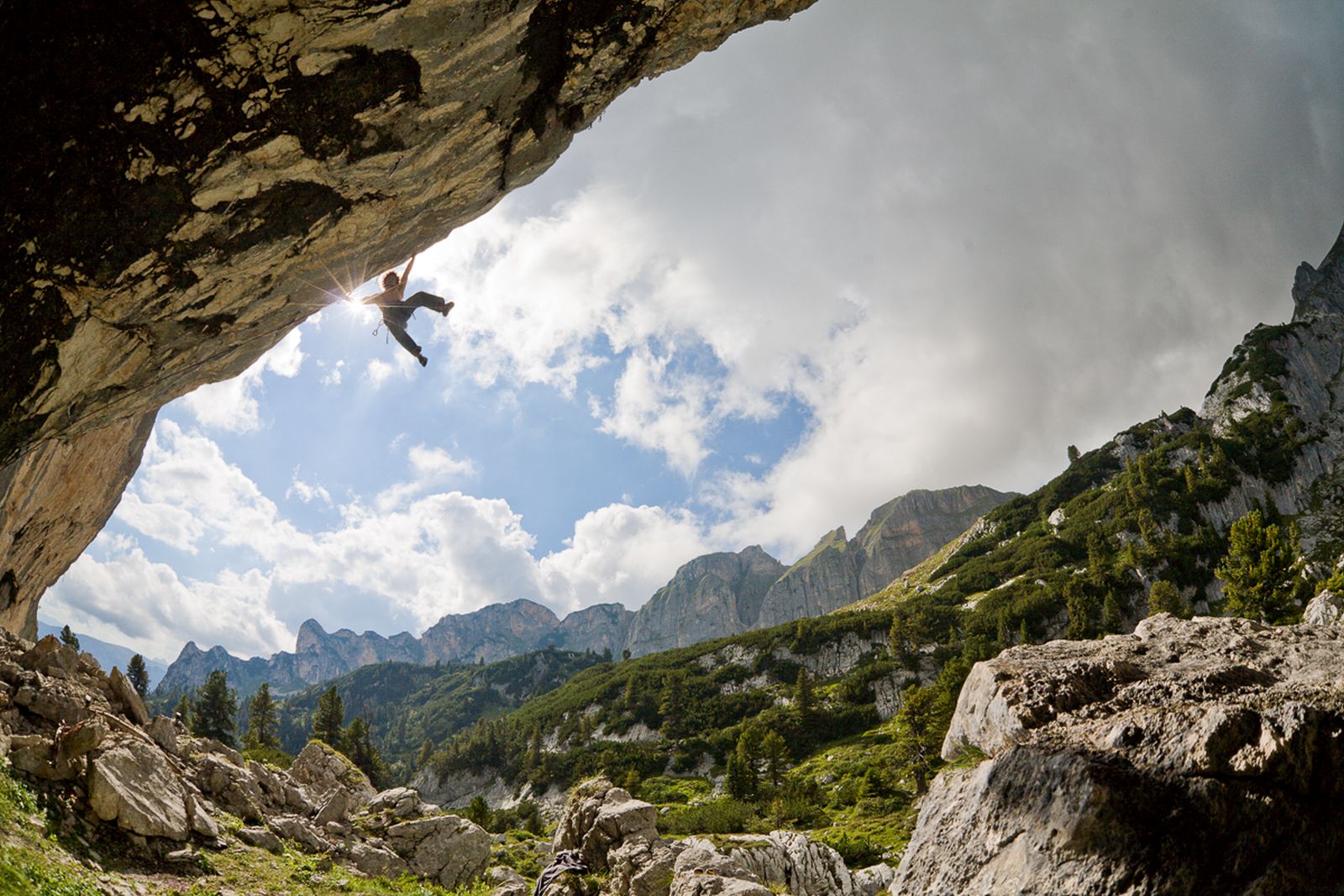 Ein Kletterer klettert aus einer Höhle an einem Seil hinauf. Im Hintergrund Berglandschaft.