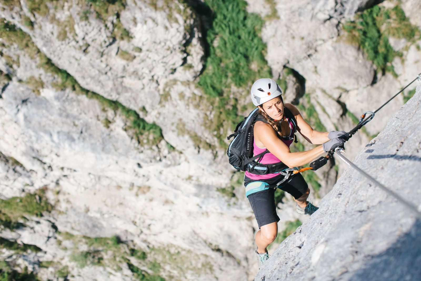 Frau klettert entlag eines Drahtseiles nach oben. Im Hintergrund hellgrauen Fels.