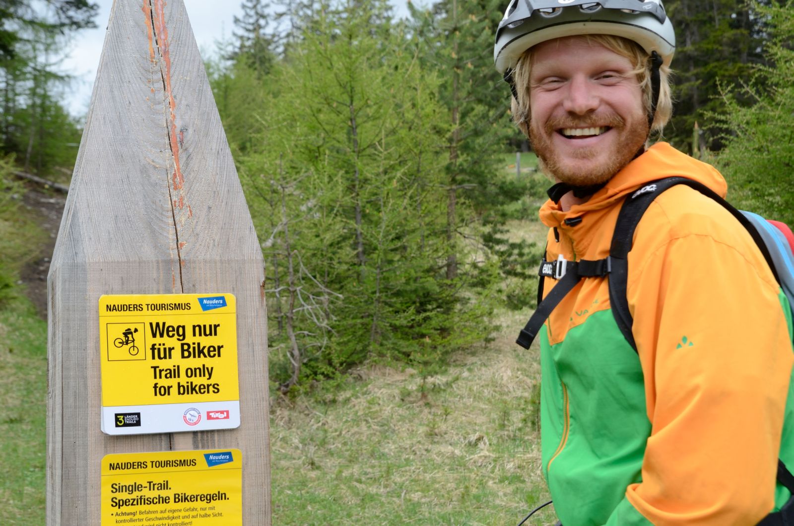 Ein Mountainbiker steht neben einem Singletrailschild und lächelt.