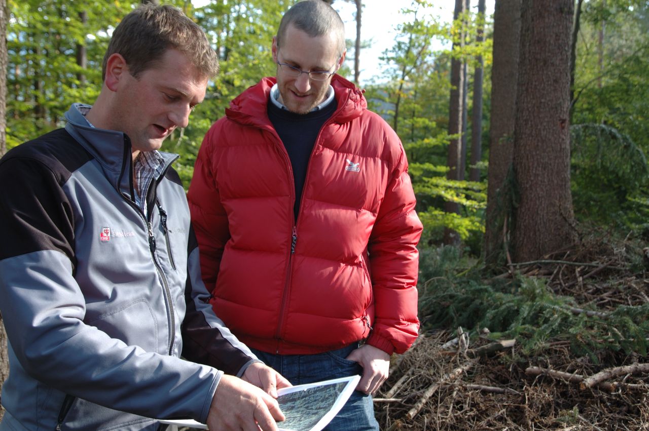 Zwei Männer schauen auf einen Plan im Wald und beraten sich.