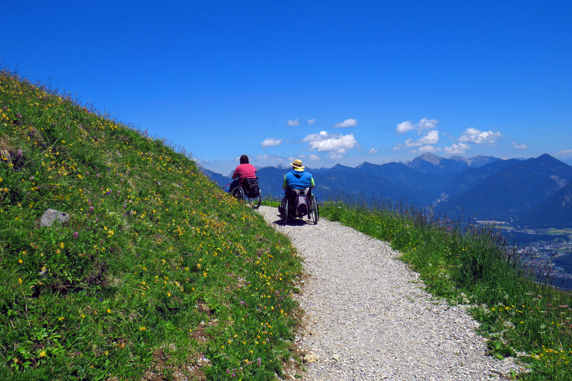 Man sieht zwei Männer im Rollstuhl sitzend von uns weg fahren. Der Spazierweg geht durch eine satt grüne Bergwiese. Im Hintergrund sieht man ins Tal beziehungsweise die Berge, über denen ein paar kleine Wolken sind. Ansonsten ist der HImmel strahlend blau. 