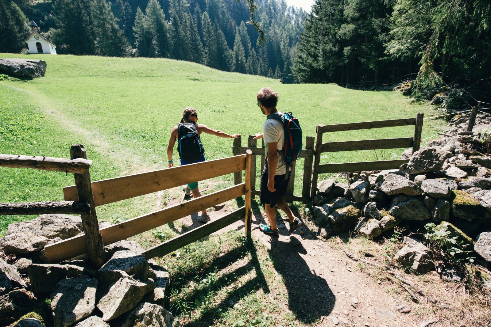 Ein Zaun und Gatter trennen einen Wiese ab. Dahinter führt ein schmaler Weg über die Wiese. Eine Frau ist durch das Gatter gegangen und hält es dem Mann auf.