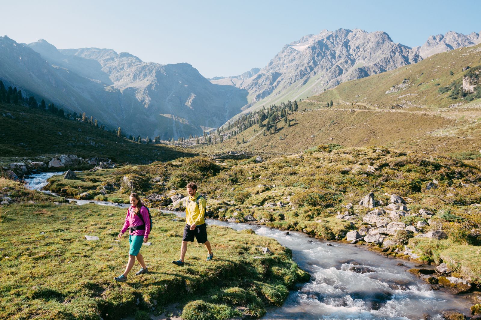 Zwei Wanderer wanderen an einem Bach entlang, welcher durch eine Almwiese fließt. Im Hintergrund Berge. 