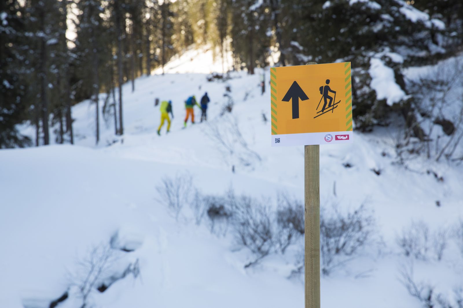 Aufstiegsschild des Skitourenleitsystems, drei Skitourengeher im Hintergrund