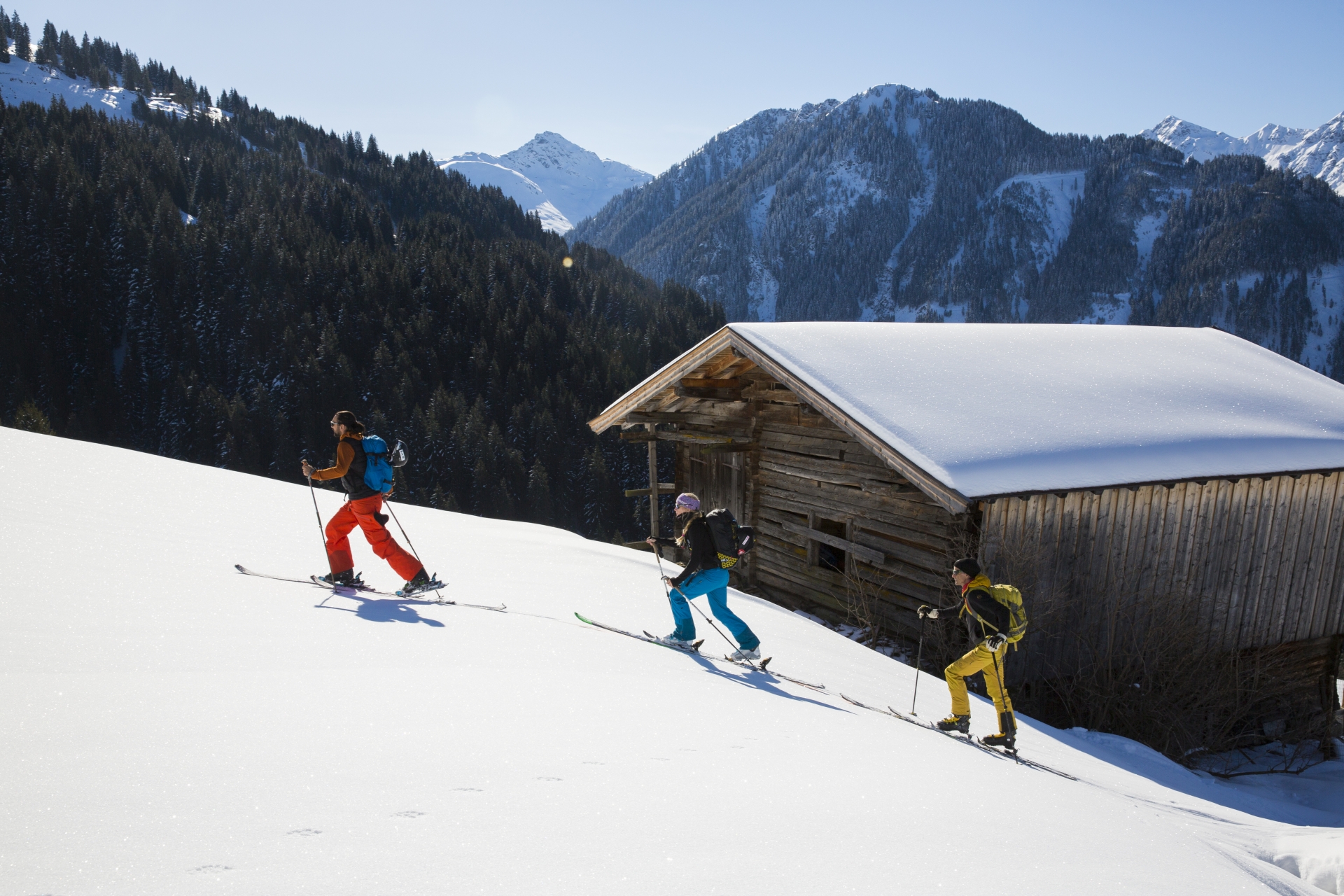 Drei Tourengeher gehen im Gelände bergauf. Im Hintergrund eine Hütte und Berge